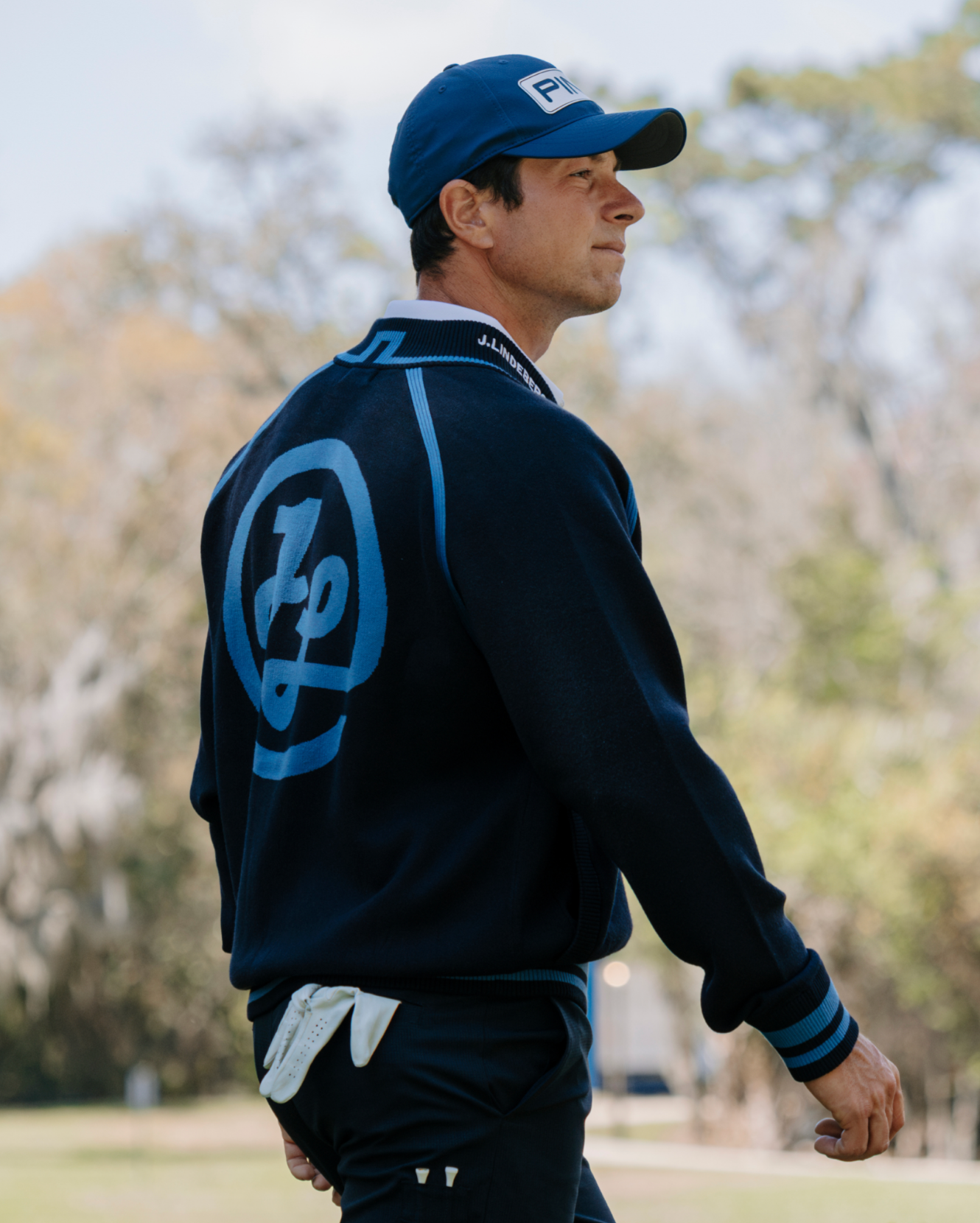 Man wearing a navy blue jacket with a logo, cap, and golf glove on a blurred outdoor background